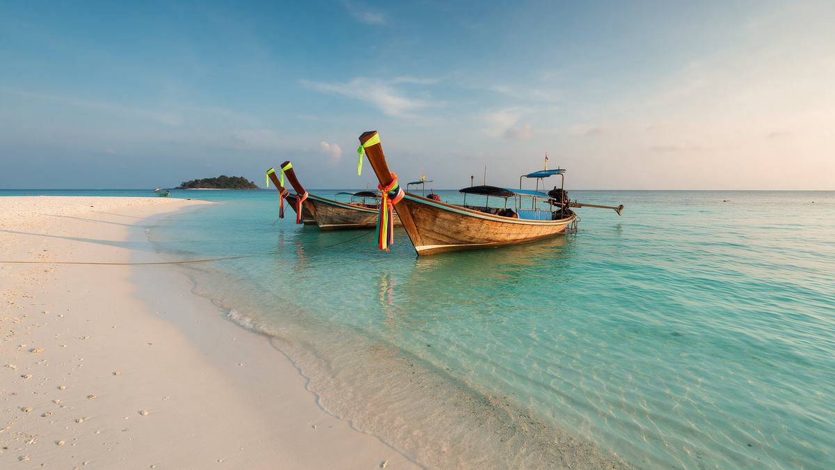 Klares türkisfarbenes Flachwasser und weißer Sandstrand auf Koh Lipe, davor Longtail-Boote mit bunten Bändern, im Hintergrund eine kleine Insel