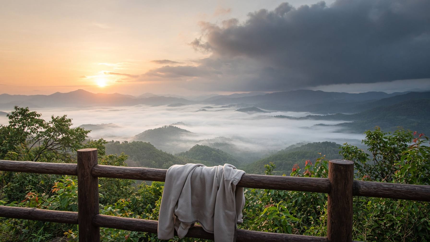 Aussichtspunkt im Norden Thailands am frühen Morgen: Nebelmeer im grünen Tal, Sonne bricht durch, dunkle Gewitterwolken über den Hügeln, eine zusammengelegte helle Jacke hängt am Holzgeländer